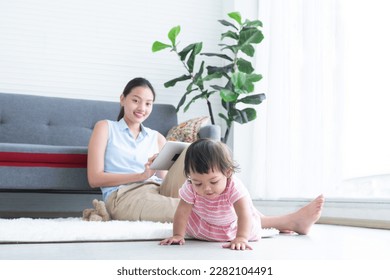 Young Asian beautiful mother sitting on floor, working with tablet while looking at her cute little Caucasian 7 months newborn baby crawling on floor playing near mom at home. Child care concept - Powered by Shutterstock