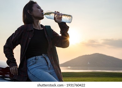 Young Asian Beautiful Tourists Relax In Front Of Their Cars And Drink Thirsty Water From Bottle With Sunlight. In Evening Atmosphere Of The Dam With Mountains And Good Weather For Relaxing And Camping