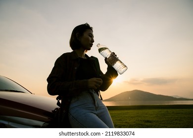 Young Asian Beautiful Tourists Relax In Front Of Their Cars And Bottle Of Water From Bottle With Sunlight. In Evening Atmosphere Of The Dam With Mountains And Good Weather For Relaxing And Camping