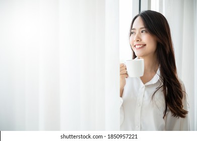 Young Asian Beautiful Woman Clothing White Pyjamas And Drink Coffee Beside Window At Morning In Bedroom. She Feeling Happy Fresh And Smile Relax