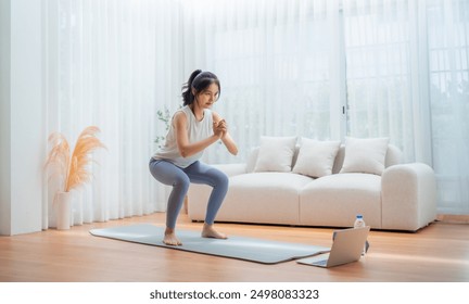 Young Asian beautiful woman doing aerobic exercise at home. Attractive girl doing  workout by squatting, follow instructions video from online trainer for health in house - Powered by Shutterstock