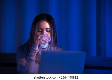 Young Asian Beautiful Woman Enjoy Using Computer Notebook To Social Online And Drink A Glass Of Milk On Bed With Happy Mood In Dark Bedroom. Insomnia Effect