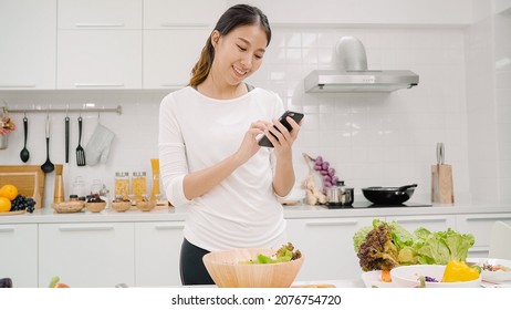 Young Asian Blogger Woman Using Smartphone Photo Post In Social Media In The Kitchen, Female Making Salad At Home. Lifestyle Women Relax At Home Concept.