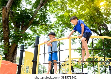 Young Asian Boy Climb On The Black Rope Fence And Yellow Bar By His Hand To Exercise At Out Door Playground Under The Big Tree.