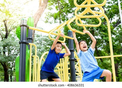 Young Asian Boy Hang The Yellow Bar By His Hand To Exercise At Out Door Playground Under The Big Tree.