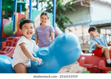 A young Asian boy, happy smile as rides a rocking horse at the playground,  his mother standing close by to watch over him and ensure his safety during playtime. - Powered by Shutterstock