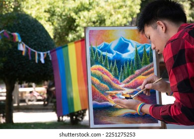 Young asian boy in plaid shirt holds paintbrush and painting his unfinished picture with a paintbrush in the park which decorated with rainbow flag and LGBTQ+ flagline, concept for genders diversity.