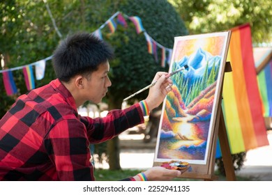 Young asian boy in plaid shirt holds paintbrush and painting his unfinished picture with a paintbrush in the park which decorated with rainbow flag and LGBTQ+ flagline, concept for genders diversity.