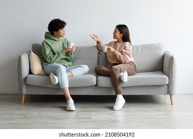 Young Asian Boyfriend And Girlfriend Drinking Coffee And Chatting While Sitting On Sofa At Home. Affectionate Millennial Couple Enjoying Hot Drink, Having Conversation, Spending Time Together