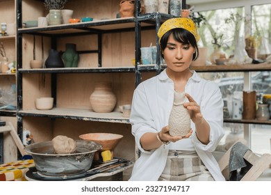 Young asian brunette female potter in headscarf and workwear holding clay sculpture near pottery wheel while working in ceramic workshop, craftsmanship in pottery making