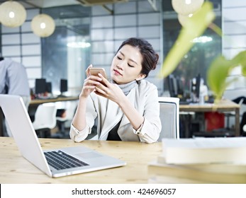 young asian business executive sitting at desk in office playing with mobile phone.