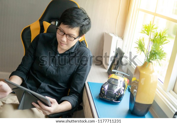 Young Asian business man\
with eyeglasses using digital tablet while sitting on working chair\
in bedroom. Home living lifestlyle with modern electronic gadget\
concept