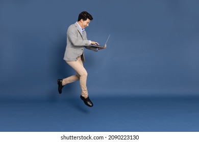 Young Asian Business Man Jumping In Air Using Laptop Computer Isolated On Deep Blue Background, Full Length Side View Concept