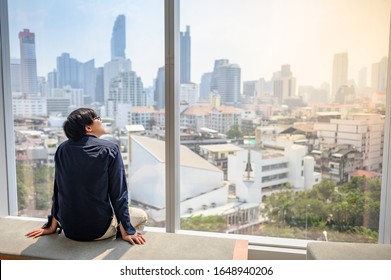 Young Asian Business Man Sitting On Bench In Office Building Looking Through The Glass Window With Cityscape Outside. Businessman Relaxing During Working In Workplace. Work Life Balance Concept