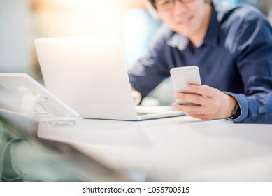 Young Asian Business Man Using Smartphone While Working With Laptop Computer In Co Working Space. Freelance Or Digital Nomad Lifestyle Concepts