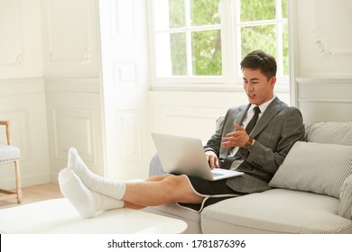 Young Asian Business Man Wearing Suit And Shorts Working From Home Meeting With Colleagues Online Using Video Chat On Laptop Computer