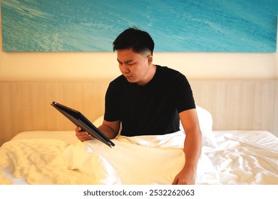 Young asian business man working on a business trip video calling his colleague and working with tablet while relaxing on bed in a luxury hotel room and this is a travel and business concept. - Powered by Shutterstock