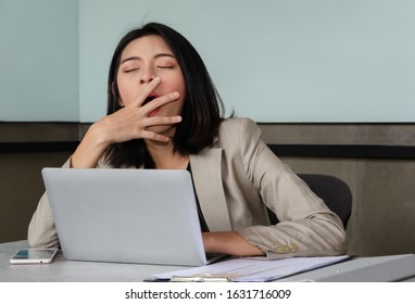 Young Asian business woman covering opened mouth with hand while yawning at meeting room in front of laptop, , feeling sleepy and bored. Overwork and sleep deprivation concept