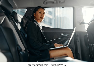 Young asian business woman with a modern laptop sitting in the back seat of luxury car. Successful startup, distance working, female boss concept.
