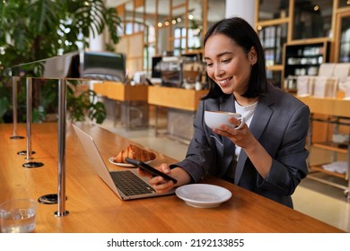 Young Asian business woman wearing suit drinking coffee using smartphone in cafe. Happy smiling female professional working holding mobile phone using smartphone texting messages on cellphone. - Shutterstock ID 2192133855