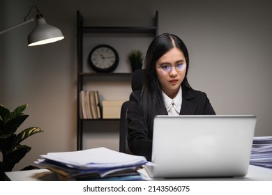Young asian business woman working at late night. She was very busy checking paperwork and had to send the work before the deadline, feeling stressed. Sitting in the dark office at night. - Shutterstock ID 2143506075