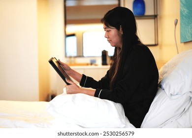 Young asian business woman working on a business trip video calling her colleague and working with tablet while relaxing on bed in a luxury hotel room and this is a travel and business concept. - Powered by Shutterstock