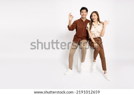 Young Asian businessman and businesswoman smiling and showing OK sign and sitting on white box isolated on white background