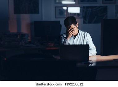 Young Asian Businessman Looking Exhausted With His Head In His Hand While Working Alone At His Desk In An Office Late At Night