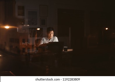 Young Asian Businessman Sitting Alone At His Desk Working On A Laptop In A Dark Office Late At Night