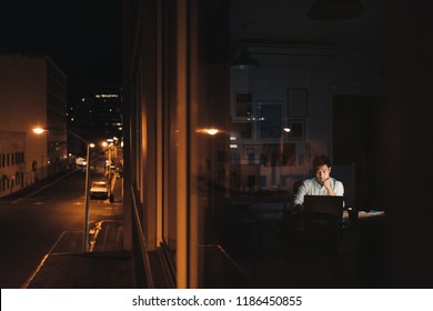 Young Asian businessman sitting at his desk working on a laptop late at night behind office building windows in the city at night