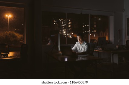 Young Asian Businessman Sitting At His Desk Working On A Laptop In A Dark Office At Night With City Lights In The Background