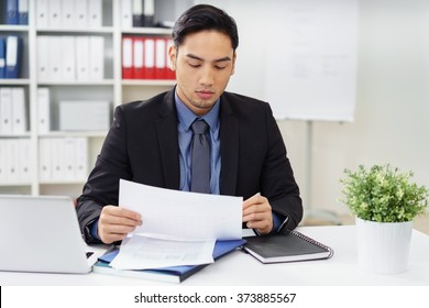 Young Asian Businessman Sitting At His Desk In The Office Proof Reading A Report Making Notes With A Pen As Her Reads Through It