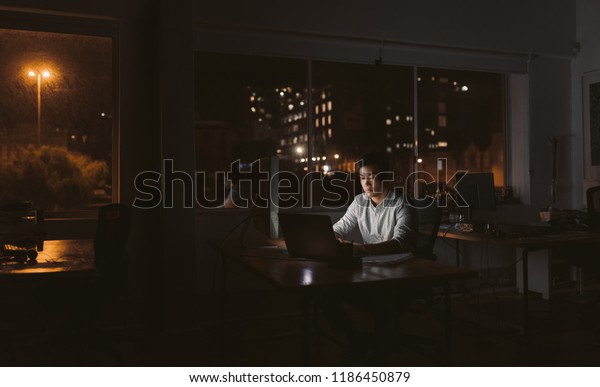 夜 暗いオフィスで 背景に街灯を持つノートパソコンを机に座って作業するアジアの若いビジネスマン の写真素材 今すぐ編集