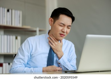 Young Asian Businessman Sitting Working With Laptop At Desk In The Office, Hand At His Neck, Feeling Unwell, Have A Sore Throat,