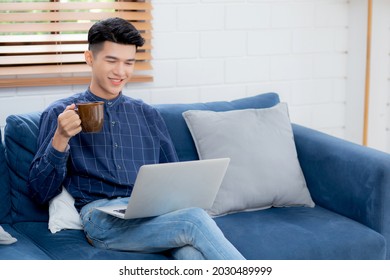 Young asian businessman smile and work from home with laptop computer online on sofa in living room, freelance business man using notebook and drink coffee on couch, new normal, lifestyle concept.