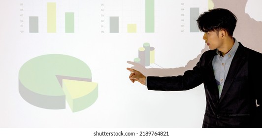 Young Asian Businessman In Suit Giving Presentation With Projector Screen. White Wall With Pie Chart And Graph. Training Staff, Meeting, Report, Business School.