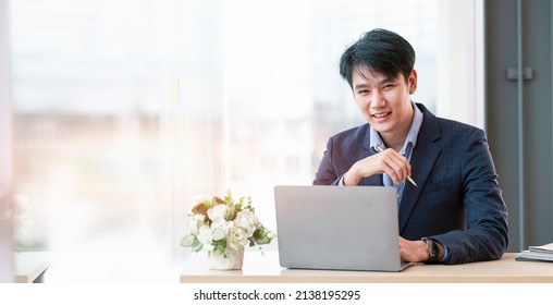 Young Asian Businessman In Suit Sitting At Desk With Laptop Computer, Smiling And Looking At Camera.