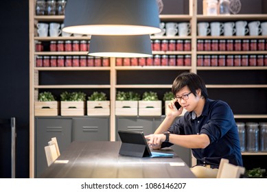 Young Asian Businessman Using Smartphone For Business Talk Pointing On His Digital Tablet In Co Working Space. Freelance Lifestyle Or Digital Nomad Concepts