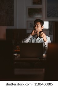 Young Asian Businessman Working At His Desk In An Office Late At Night Yawning While Drinking A Cup Of Coffee To Stay Awake