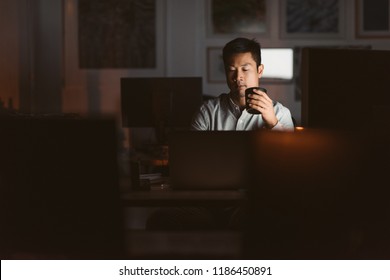 Young Asian businessman working at his desk in an office late at night drinking a cup of coffee to stay awake - Powered by Shutterstock