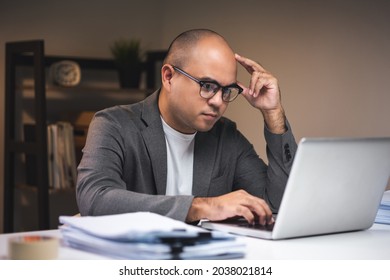 Young Asian Businessman Working With Laptop Computer In The Dark Office At Night. Attractive Indian Man Work Hard Overtime Serious Thinking In Home With Floor Lamp Ambient Warm Light Late At Night