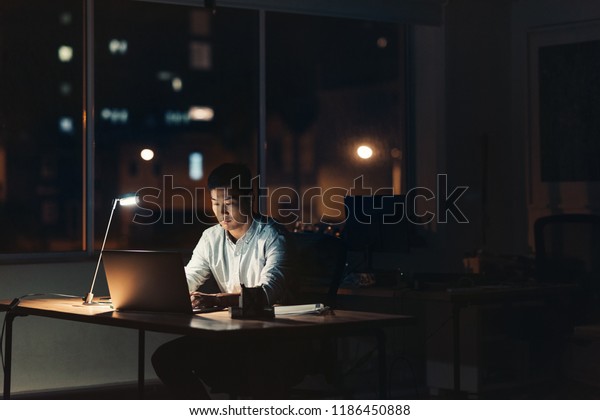 夜 暗いオフィスの机に座りながら 背景に街灯を持つノートパソコンを使用するアジア系の若いビジネスマン の写真素材 今すぐ編集