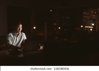 Young Asian Businessman Working Overtime Alone At His Desk In An Office Late At Night With City Lights Glowing In The Background