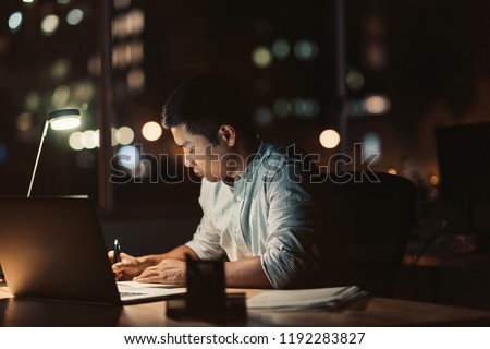 Young Asian businessman writing in a notebook while working at his desk in a dark office at night with city lights in the background