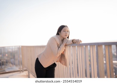 Young asian businesswoman in a beige blazer and black pants leaning against the railing of a rooftop at sunset, contemplating the stunning cityscape and feeling inspired by the moment - Powered by Shutterstock