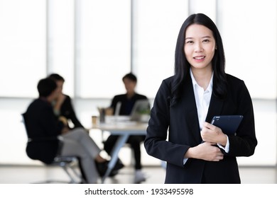 Young Asian Businesswoman In Black Suit Standing And Holding Tablet  Computer Posing To Camera In Office With Her Team Talking In Blur Background.