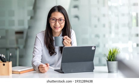 Young Asian businesswoman Looking at the camera, drinking coffee, working on a tablet at the office.