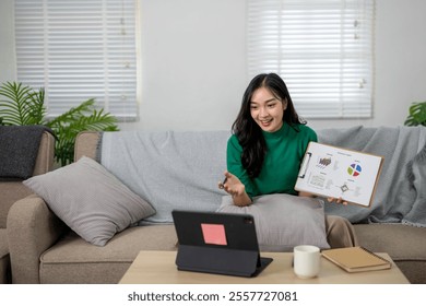 Young asian businesswoman is making a video call while sitting on a sofa, holding a financial report, and gesturing with her hand - Powered by Shutterstock