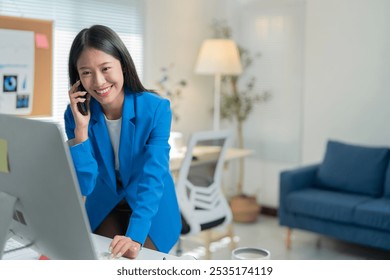 Young asian businesswoman multitasks happily in a modern office, talking on her phone and working on her computer, exuding confidence and professionalism - Powered by Shutterstock