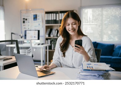 Young asian businesswoman multitasks with a smartphone and laptop in a modern office, focusing on data analysis, marketing, finance, and accounting - Powered by Shutterstock